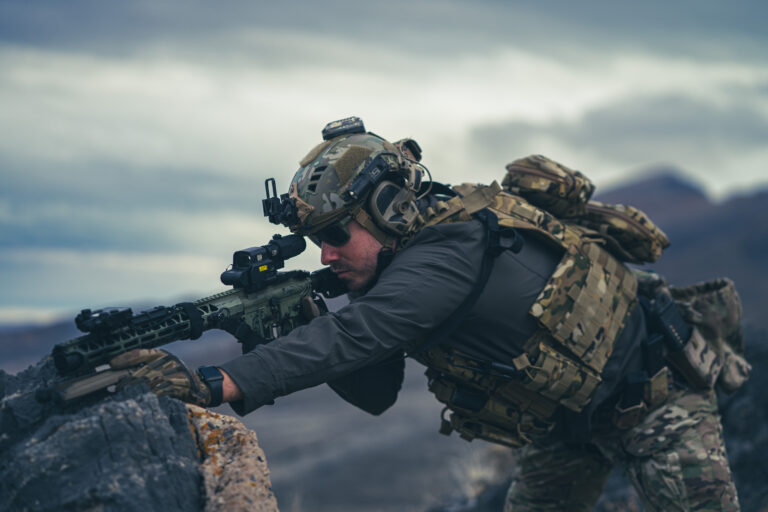 A man in camouflage protective body armor and FAST helmet sets sights on target.